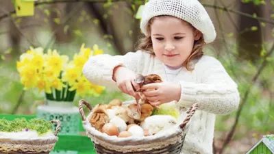 Malá dívka v bílé čepici a svetru sbírá houby do proutěného košíku v jarní zahradě s květinami a budkami v pozadí